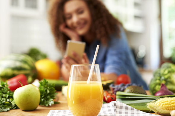 Glas Orangensaft inmitten von frischem Obst und Gemüse, im Hintergrund eine Frau mit Smartphone