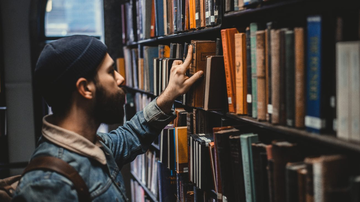 Mann in Bibliothek greift nach einem Buch im Bücherregal