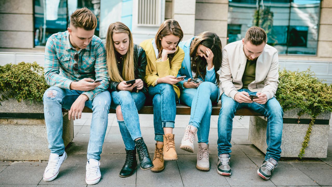 Fünf Jugendliche sitzen im Freien nebeneinander auf einer Bank und betrachten ihre Smartphones.