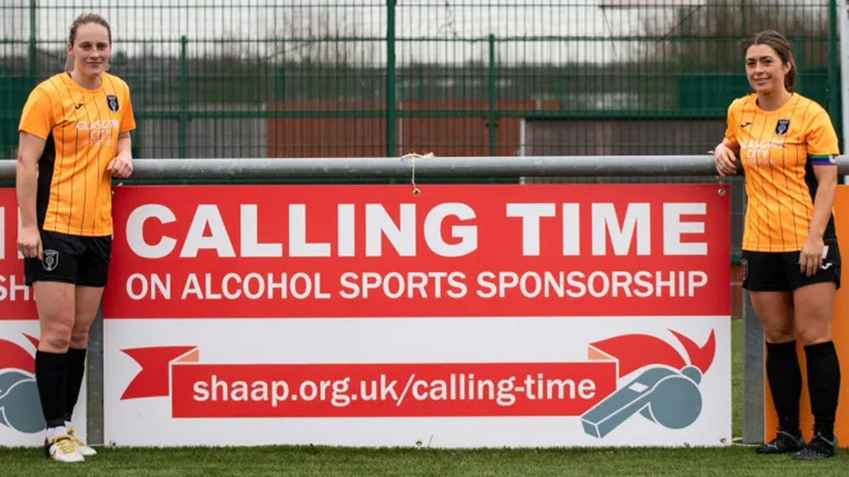 Zwei Fußballspielerinnen stehen neben einer Bandenwerbung für 'Calling Time on Alcohol Sports Sponsorship'