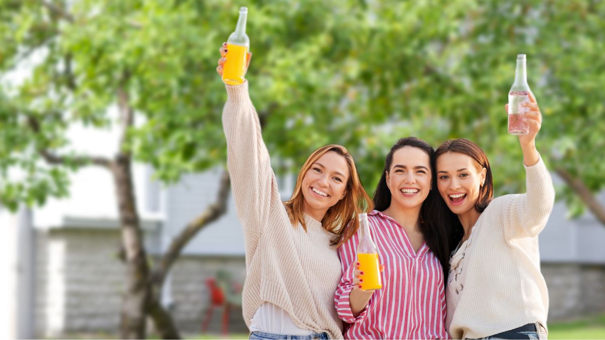 Drei Frauen prosten der Kamera mit Orangensaft- und Mineralwasserflaschen zu.