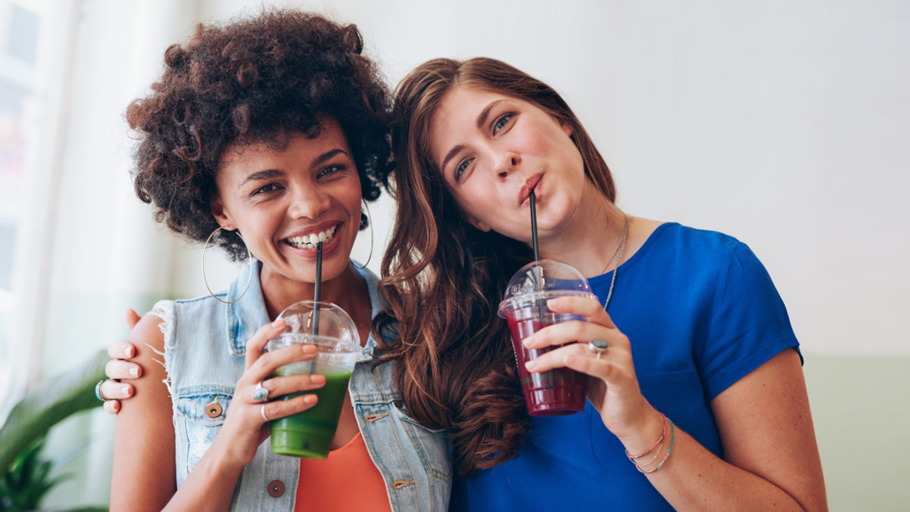 Zwei Frauen stehen nebeneinander in Umarmung und trinken frisch gepresste Fruchtsäfte.