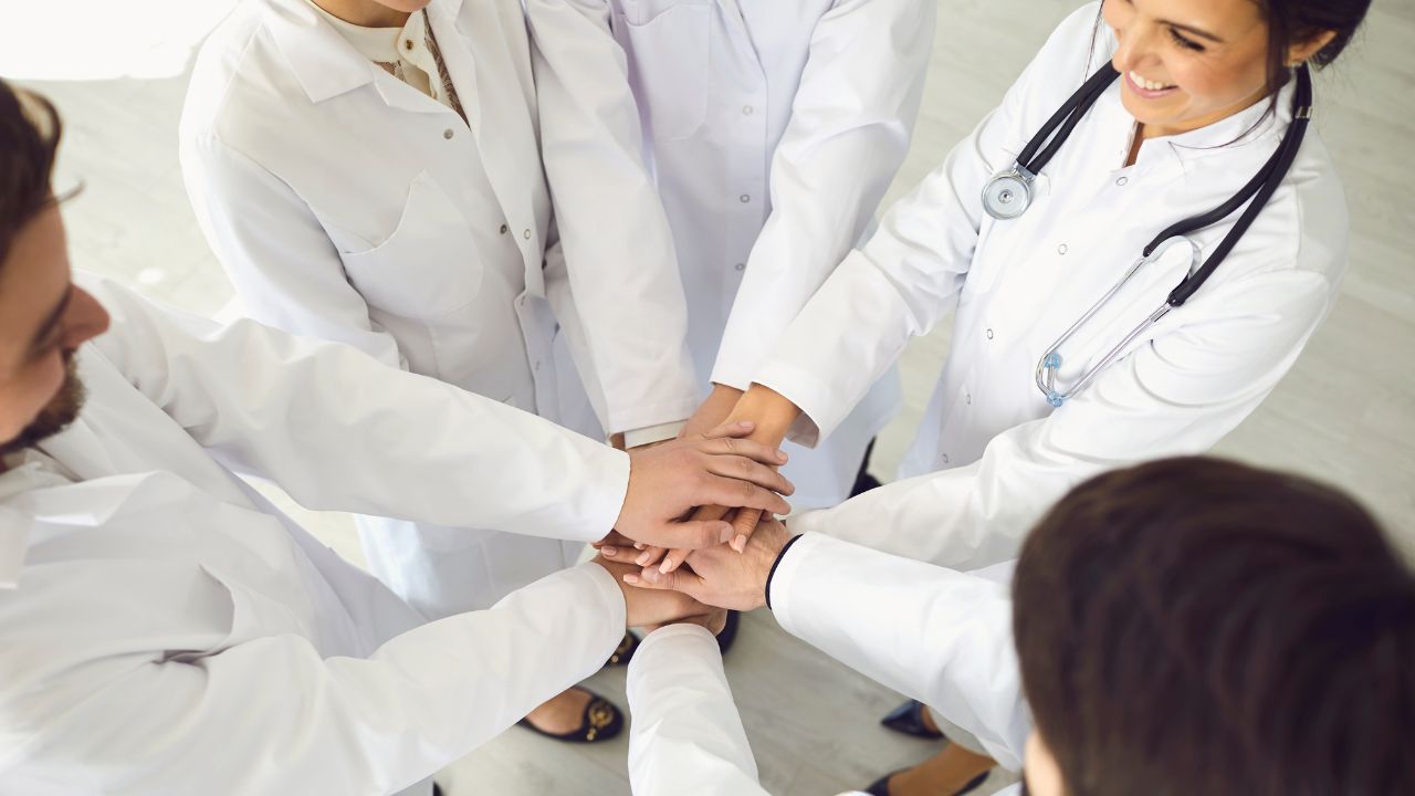 Mehrere Ärztinnen und Ärzte in weißen Kitteln stehen im Kreis und legen ihre Hände übereinander als Zeichen für Teamwork und Zusammenarbeit.