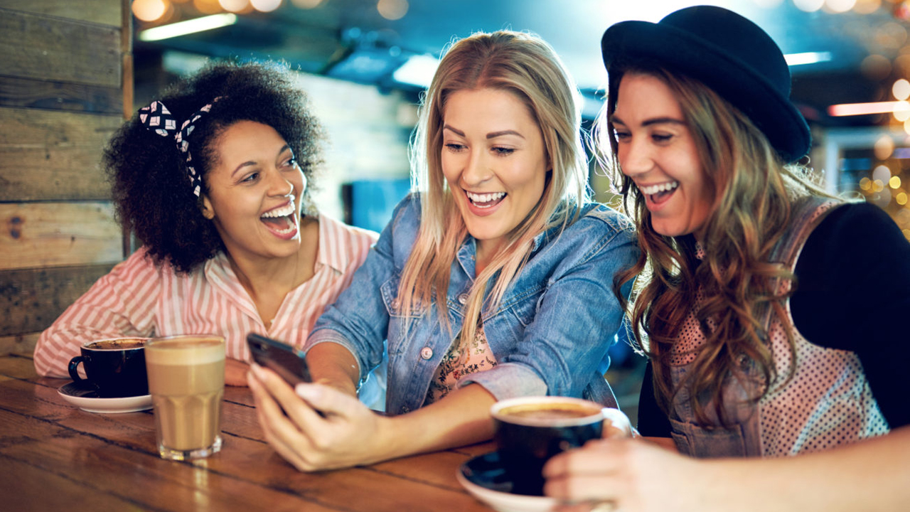 Drei junge Frauen in Kaffeebar betrachten lachend ein Smartphone.