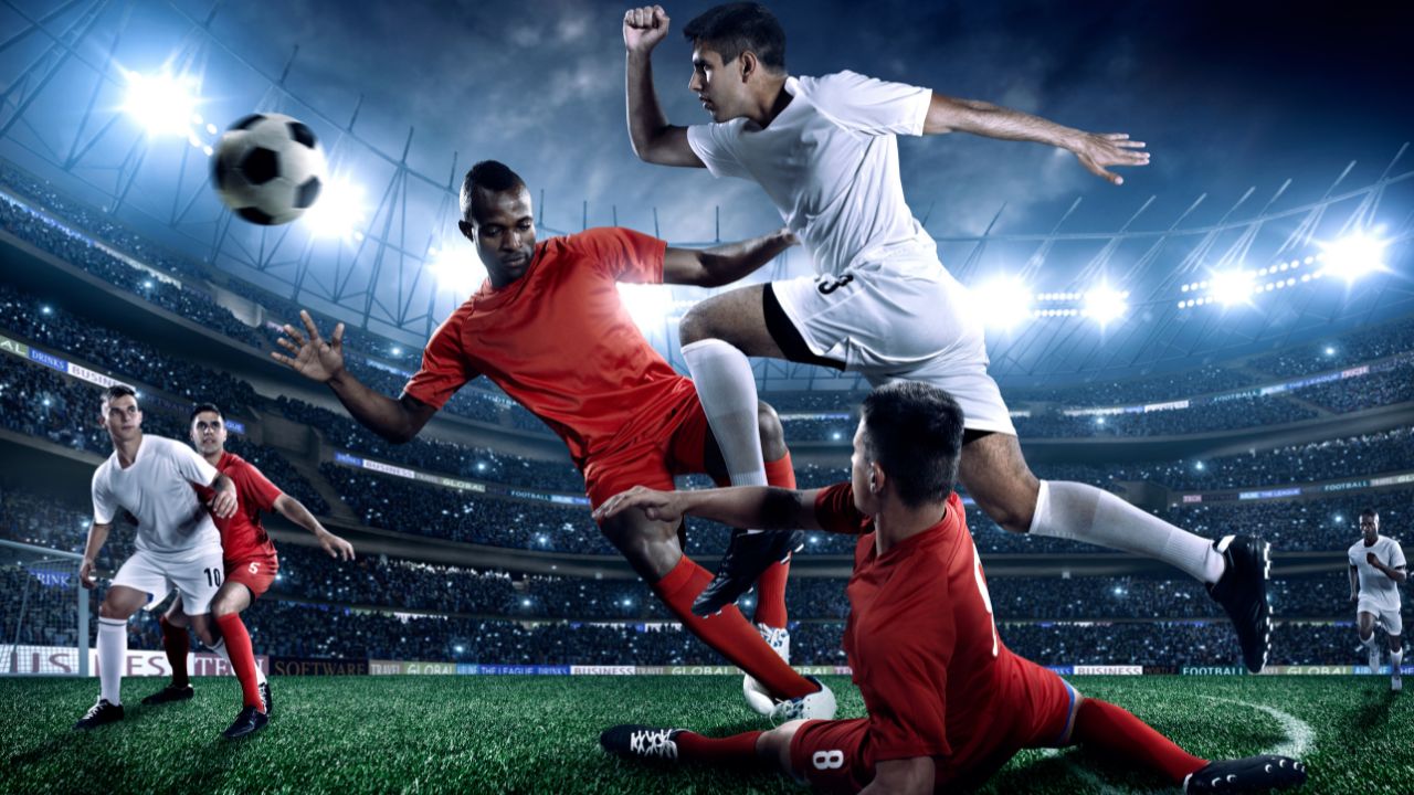 Ein Stürmer in weißer Fußballkleidung springt über zwei Verteidiger in roter Fußballkleidung und jagt dem Ball hinterher.