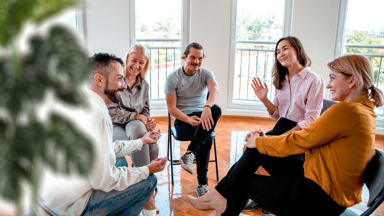 Dieses Bild zeigt eine Gruppe von fünf Personen, die in einem hellen, luftigen Raum mit großen Fenstern sitzen und sich unterhalten. Die Personen sind in einem Kreis angeordnet, was auf eine informelle oder therapeutische Gruppensitzung hinweist. Sie wirken entspannt und engagiert, einige lächeln und gestikulieren, was auf eine positive und unterstützende Atmosphäre hindeutet. Der Raum ist mit modernen Möbeln und Pflanzen dekoriert, was zur einladenden und ruhigen Umgebung beiträgt.