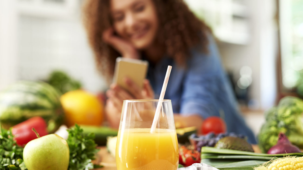 Ein Glas Orangensaft auf einem mit Gemüse bedeckten Tisch, an dessen Ende im Hintergrund unscharf eine Frau zu erkennen ist, die lächelnd ihr Smartphone betrachtet.