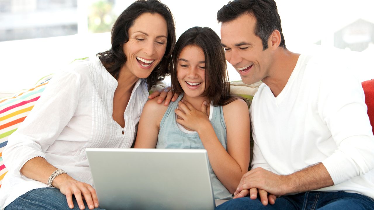 Jugendliches Mädchen sitzt zwischen seinen Eltern auf der Couch. Alle betrachten lachend den Bildschirm eines Laptops.