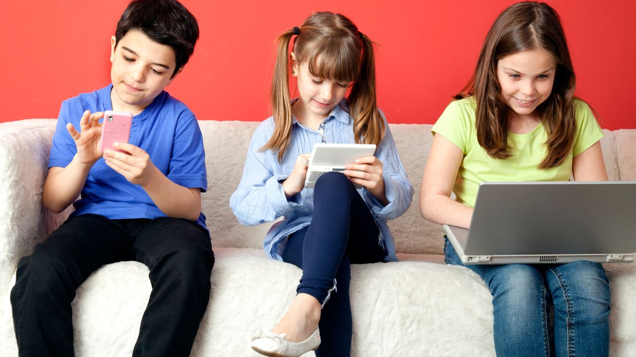 Drei Kinder sitzen auf einem Sofa und beschäftigen sich mit ihren Smartphones, Tablets und Laptops.