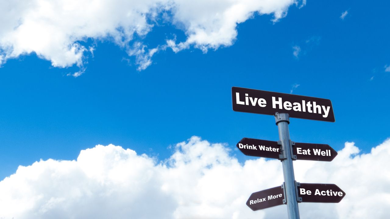 Das Bild zeigt ein Schild mit verschiedenen Gesundheitshinweisen vor einem blauen Himmel mit weißen Wolken. Auf dem braunen Schild stehen die englischen Aufforderungen 'Live Healthy', 'Drink Water', 'Eat Well', 'Relax More' und 'Be Active'. Das Schild soll dazu motivieren, einen gesunden Lebensstil zu pflegen, indem man Wasser trinkt, sich gesund ernährt, entspannt und aktiv ist.