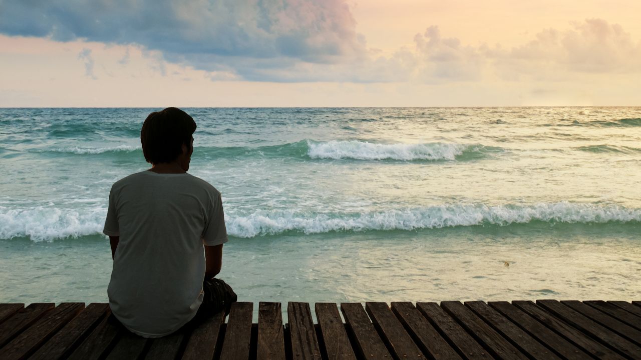 Ein Mann sitzt einsam auf einem Holzsteg am Ufer und blickt aufs Meer hinaus.