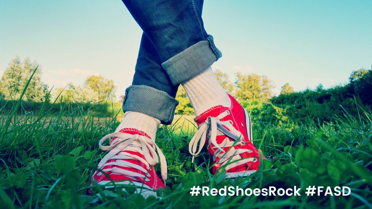 Person in roten Turnschuhen mit niedrigem Schaft steht mit gekreuzten Beinen auf Gras. Darunter die Hashtags 'Red Shoes Rock' und 'FASD'.