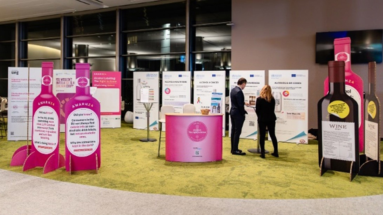 Messestand im Hauptgebäude des Europäischen Parlaments in Brüssel mit Informationsmaterialien und Aufstellern in Form von Weinflaschen. Zwei Personen betrachten Plakate zur Alkoholaufklärung und Kennzeichnung.