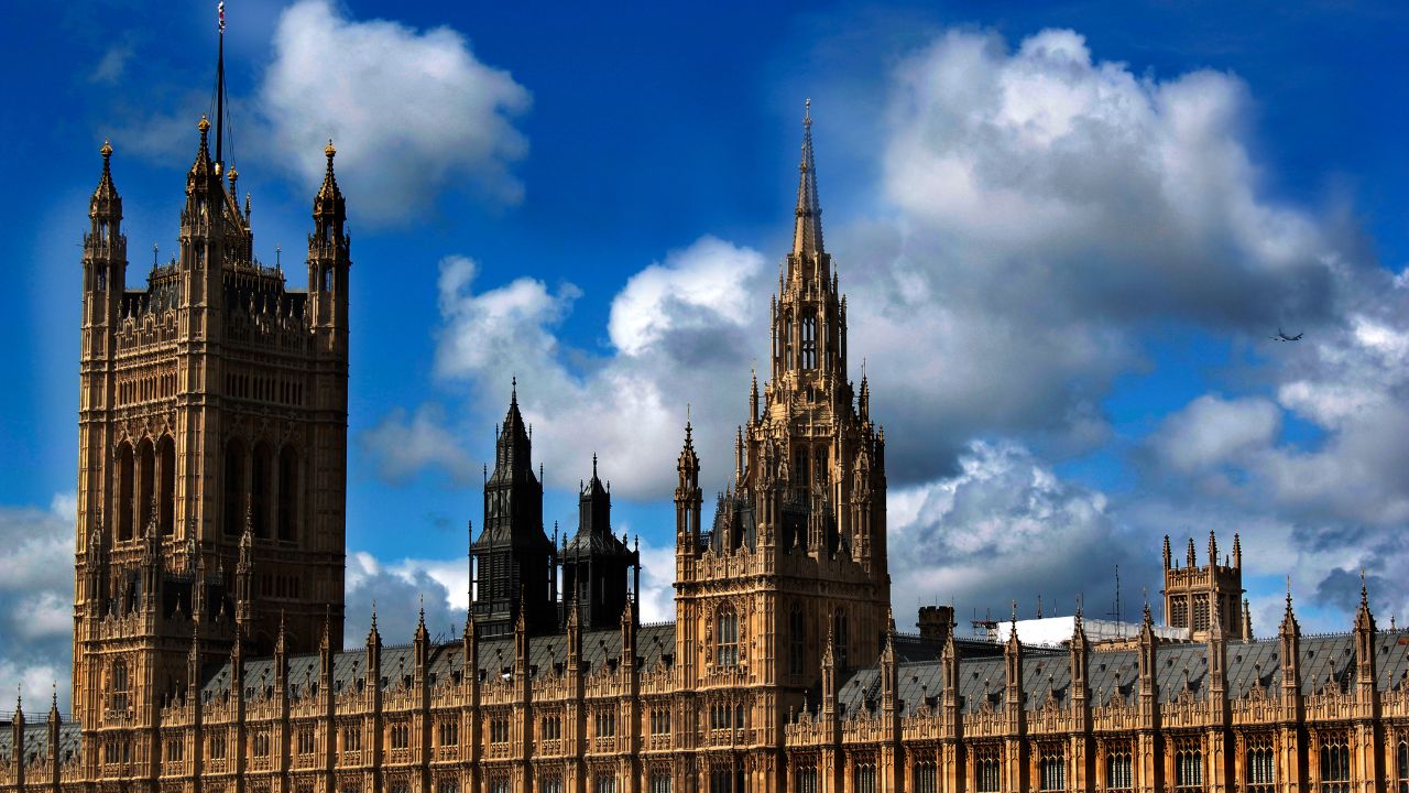 Ansicht der Houses of Parliament in London, mit dem markanten Victoria Tower links und einem schlanken, gotischen Spitzturm rechts, vor einem leuchtend blauen Himmel mit weißen und grauen Wolken. Ein kleines Flugzeug ist im oberen rechten Bereich erkennbar.
