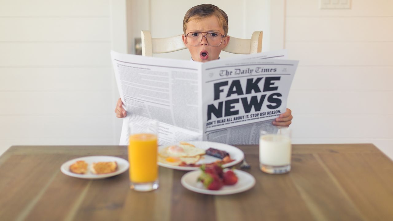 Ein junger Junge mit Brille sitzt am Frühstückstisch und blickt mit offenem Mund schockiert auf eine Zeitung. Die große Schlagzeile der Zeitung lautet 'FAKE NEWS', darunter der Text 'DON'T READ ALL ABOUT IT. STOP USE REASON.'. Auf dem Tisch stehen Frühstücksartikel wie Orangensaft, Milch und Essen.