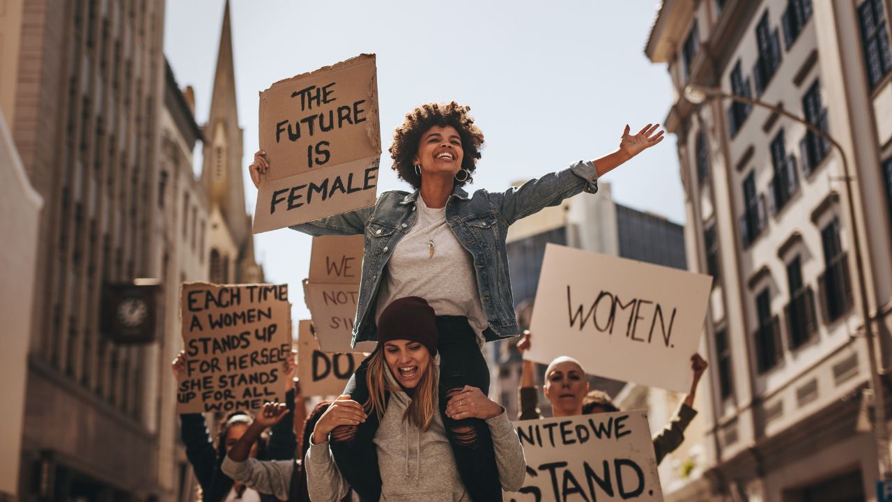 Gruppe von Frauen, die in einer Stadt an einer Demonstration teilnehmen; eine Frau sitzt auf den Schultern einer anderen und hält ein Schild mit der Aufschrift ‚The future is female‘, während weitere Teilnehmerinnen unterschiedliche Protestplakate hochhalten.