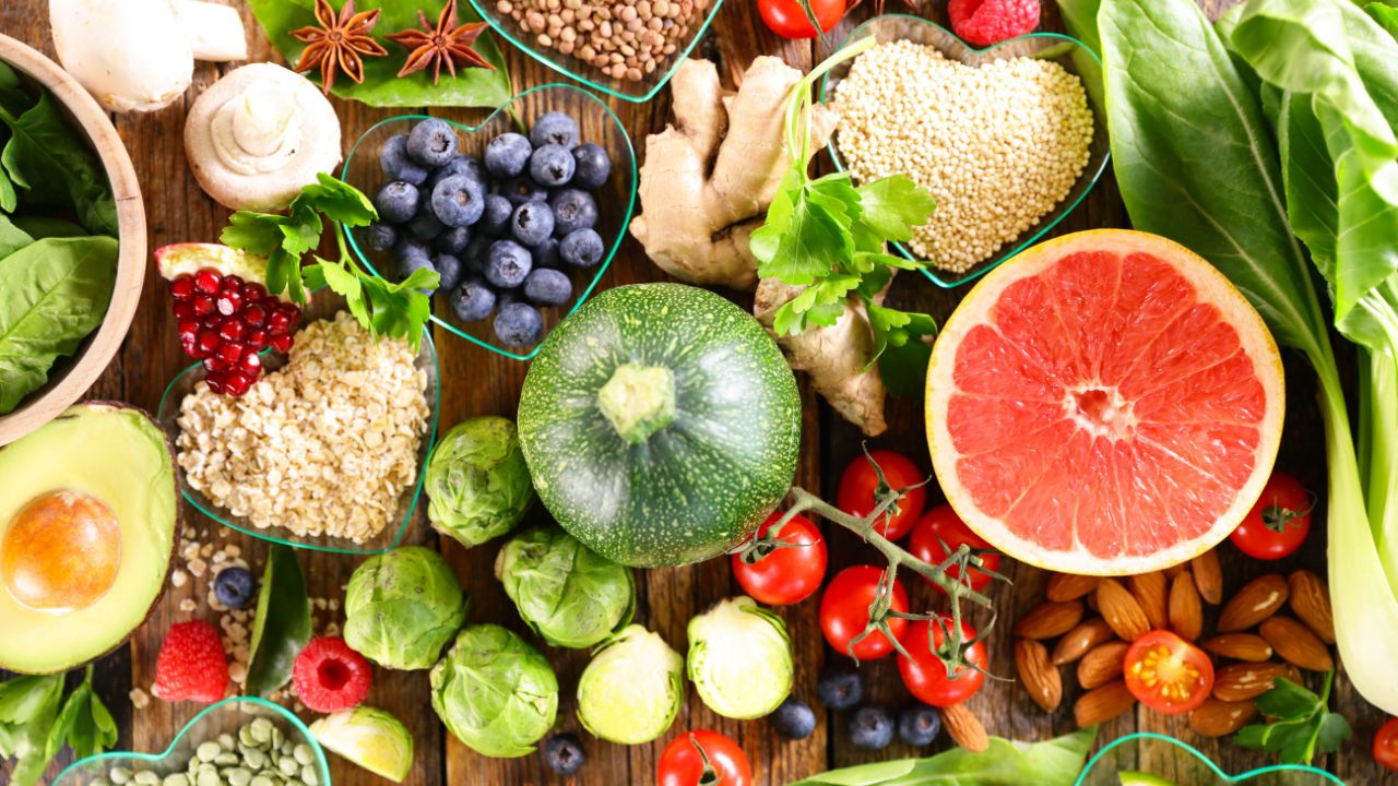 Eine bunte Ansammlung frischer, gesunder Lebensmittel auf einer rustikalen Holzoberfläche: darunter eine halbierte Grapefruit, eine Zucchini, Brokkoli, Kirschtomaten, Blaubeeren, Himbeeren, Granatapfelkerne, Avocado, Pilze, Ingwer, Haferflocken, Quinoa, Linsen, Mandeln und grünes Blattgemüse – alles kunstvoll arrangiert und von oben fotografiert.