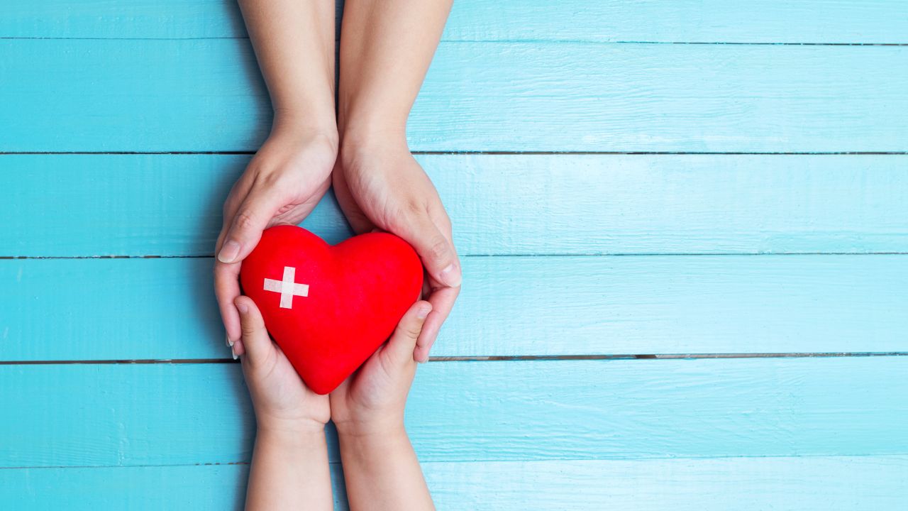Zwei Hände halten ein rotes Herz mit einem weißen Kreuz darauf, das auf einer hellblauen Holzoberfläche platziert ist. Die Szene vermittelt eine Botschaft der Fürsorge, Gesundheit und Unterstützung.