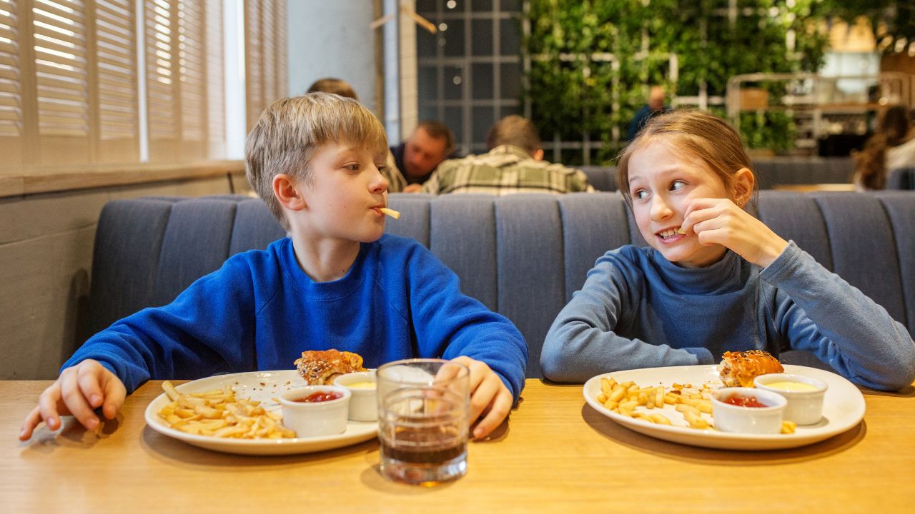 Ein Junge und ein Mädchen sitzen in einem Restaurant nebeneinander in einer Sitznische, essen lachend Pommes Frites und Burger.