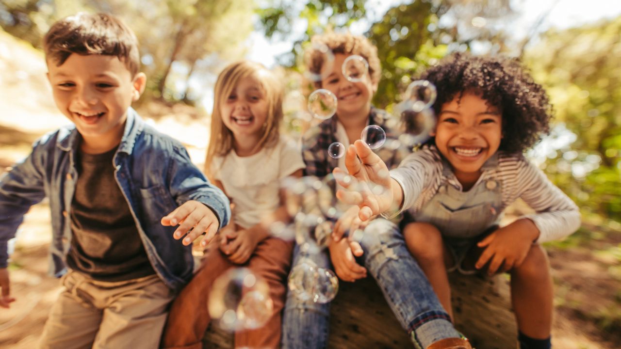 Vier lächelnde Kinder im Freien, die gemeinsam Seifenblasen machen und sich dabei sichtbar amüsieren. Die Kinder sind in lockerer Kleidung und sitzen oder knien auf einem natürlichen Untergrund, umgeben von Bäumen und Grünflächen.