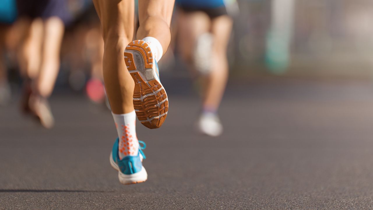 Rückansicht der Beine eines Läufers bei einem Marathon, mit Fokus auf die orangefarbene Sohle des Laufschuhs, der sich gerade vom Boden abhebt.