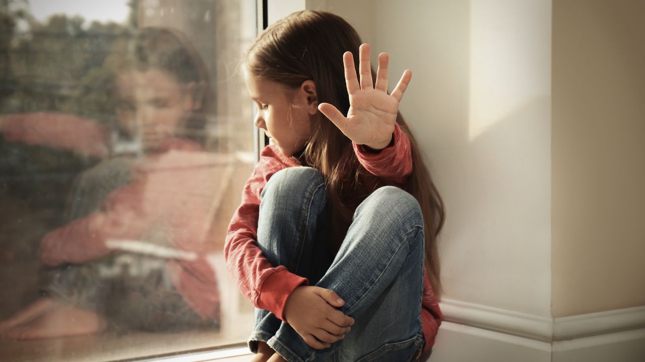 Mädchen im Jeans und rotem Pullover sitzt zusammengesunken am Fensterbrett, blickt traurig zur Seite und hält eine Hand abwehrend erhoben.
