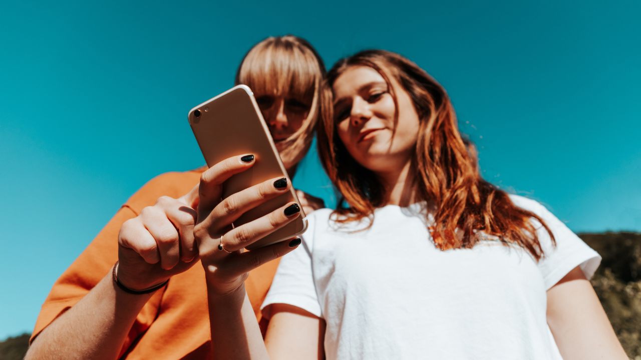 Zwei junge Frauen stehen nebeneinander und lesen gemeinsam die Beiträge in den sozialen Medien auf einem Mobiltelefon. Aufnahme von unten vor sommerlichem Himmel.