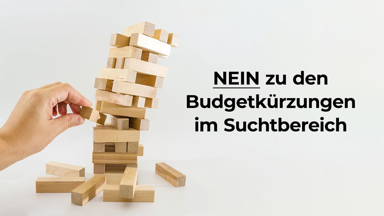 Eine Hand zieht vorsichtig einen Holzklotz aus einem hohen, instabilen Turm aus hellen Holzklötzen (ähnlich einem Jenga-Spiel), der bereits gefährlich schief steht und unten teilweise zusammengebrochen ist. Rechts im Bild steht in großer schwarzer Schrift auf weißem Hintergrund der Text: 'NEIN zu den Budgetkürzungen im Suchtbereich'. Das Bild dient als Metapher dafür, dass finanzielle Kürzungen das Hilfssystem im Suchtbereich destabilisieren und zum Einsturz bringen könnten.
