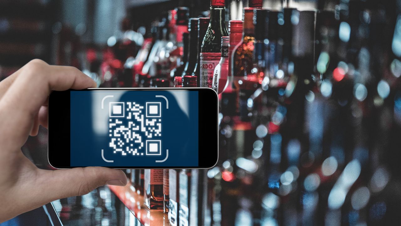 Eine Hand hält ein Smartphone mit einem QR-Code auf dem Bildschirm vor einer Auswahl an Flaschen im Hintergrund.