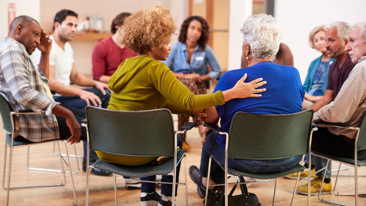 Eine diverse Gruppe von Menschen verschiedenen Alters sitzt in einem Kreis in einer Selbsthilfegruppe, wobei eine Frau einer anderen tröstend die Hand auf die Schulter legt.