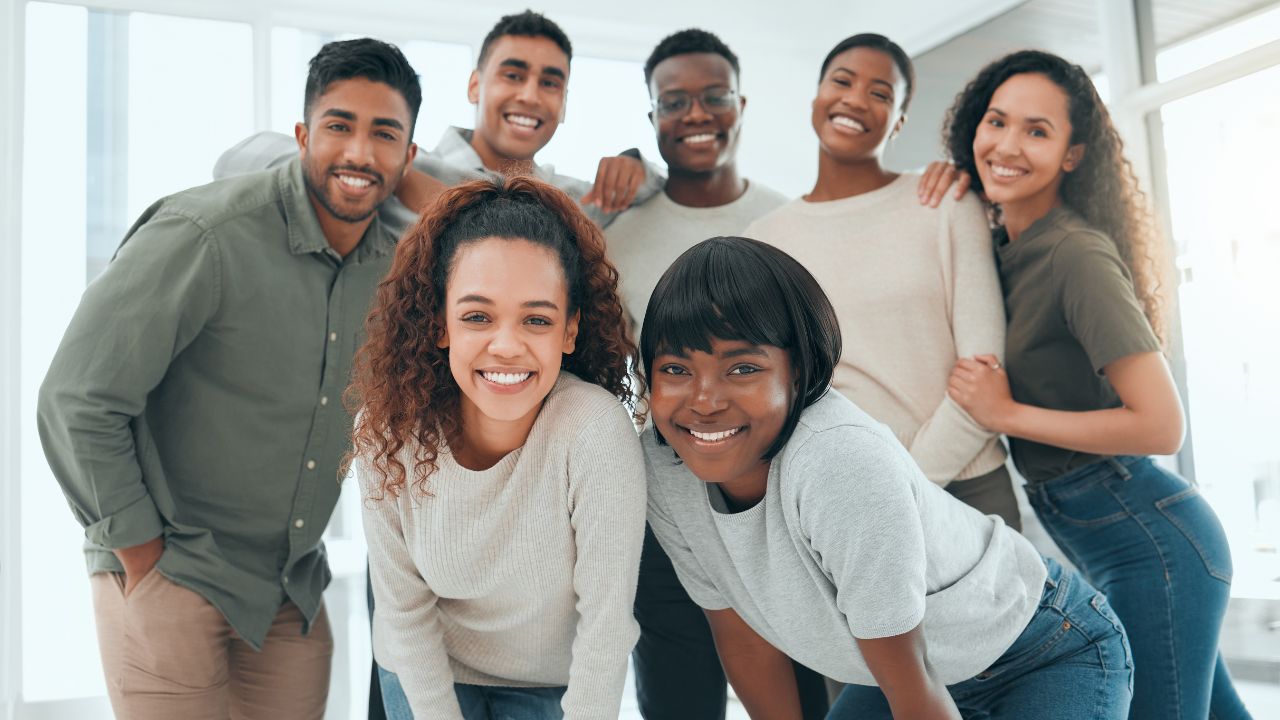 Eine Gruppe von sieben glücklichen und lässig gekleideten jungen Erwachsenen, die sich in einem modernen, hellen Raum mit großen Fenstern versammelt haben. Sie lächeln und wirken entspannt, wobei einige von ihnen einander umarmen oder die Arme umeinander legen. Die Gruppe besteht aus verschiedenen Personen, die unterschiedliche ethnische Hintergründe haben, und sie wirken wie eine freundschaftliche oder kollegiale Gruppe, die sich gut versteht. Die Atmosphäre wirkt positiv und einladend.