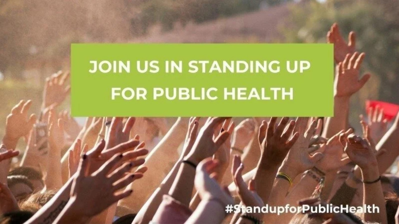 Viele erhobene Hände einer Menschenmenge im Sonnenlicht, darüber ein grünes Banner mit weißer Aufschrift 'Join us in standing up for public health' und am unteren Bildrand der Hashtag #StandupforPublicHealth.