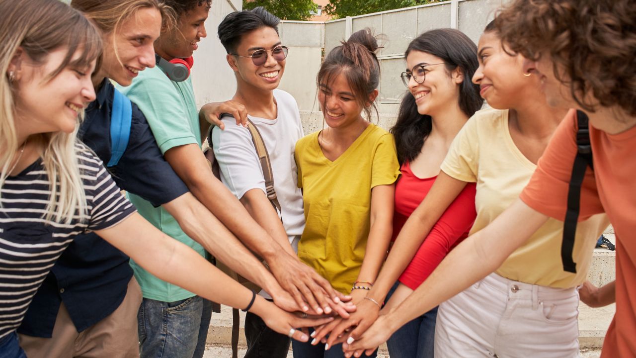 Eine Gruppe von etwa sieben jungen Erwachsenen mit unterschiedlichem Hintergrund steht eng beieinander und legt ihre Hände in der Mitte übereinander in einer Geste der Einigkeit und des Teamgeists. Sie lächeln und sehen glücklich aus.
