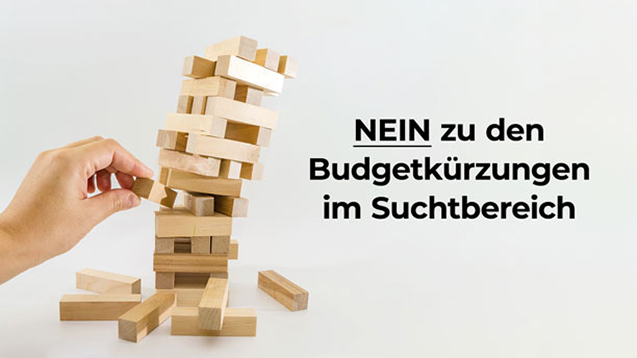 Stürzender Turm aus Holzklötzen, aus dessen mittlerem Bereich einen tragenden Stein entfernt. Dazu der Text: NEIN zu den Budgetkürzungen im Suchtbereich.