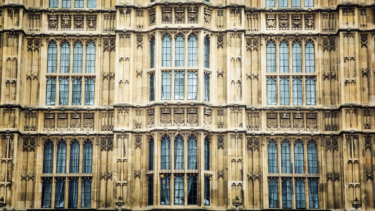 Detaillierte Ansicht der neugotischen Fassade des Palace of Westminster in London. Die streng symmetrische Fassade aus verwittertem hellem Sandstein ist über mehrere Stockwerke hinweg aus wiederkehrenden Gruppen von vier spitzbogigen Fenstern aufgebaut. Dazwischen befinden sich aufwändige steinerne Verzierungen und Wappen. Ein vorspringender Erker bildet das Zentrum der Aufnahme. Hinter fast allen Fenstern sind helle Vorhänge zu sehen, ein einzelnes Fenster ist von innen beleuchtet.