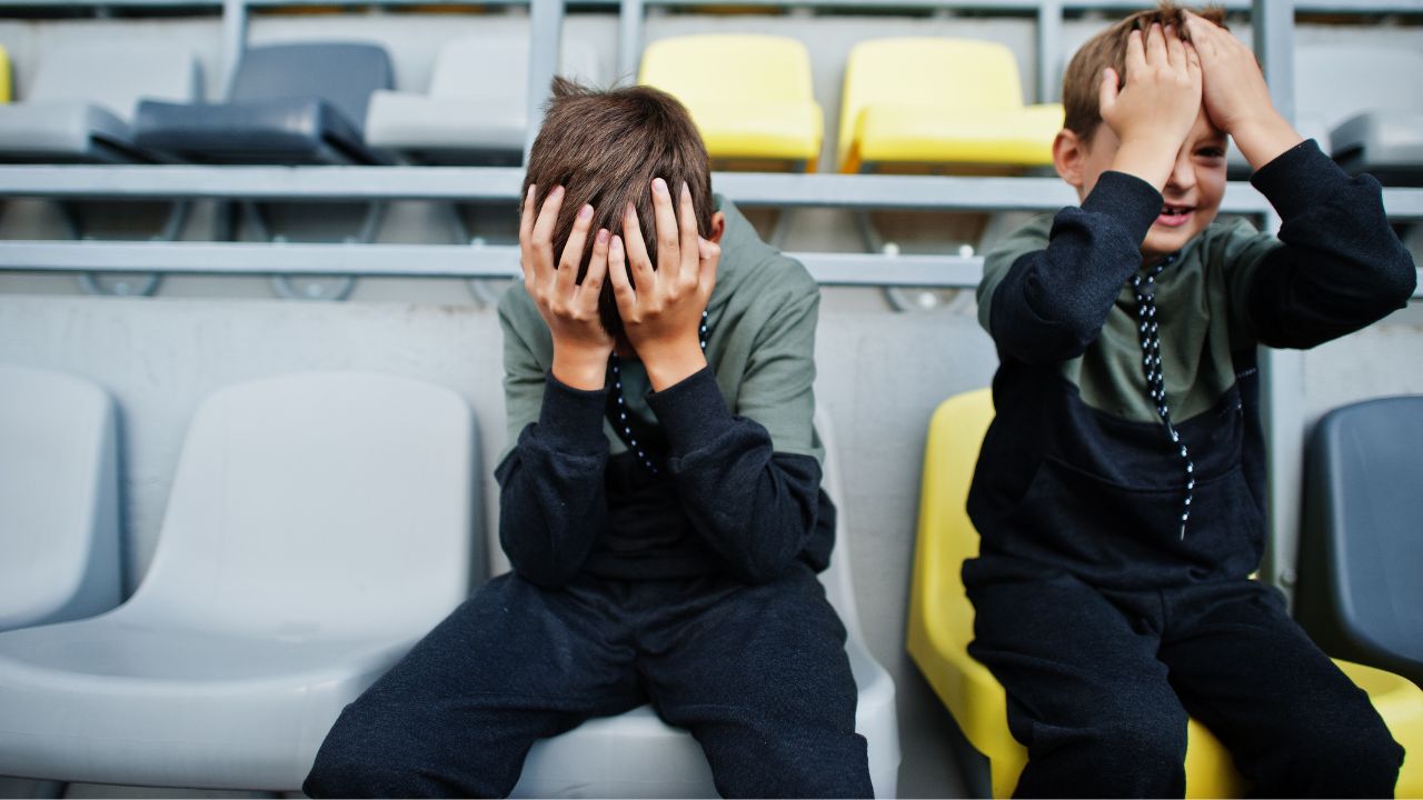Zwei enttäuschte Jungen sitzen mit den Händen vor dem Gesicht auf Tribünenplätzen in einem Stadion.