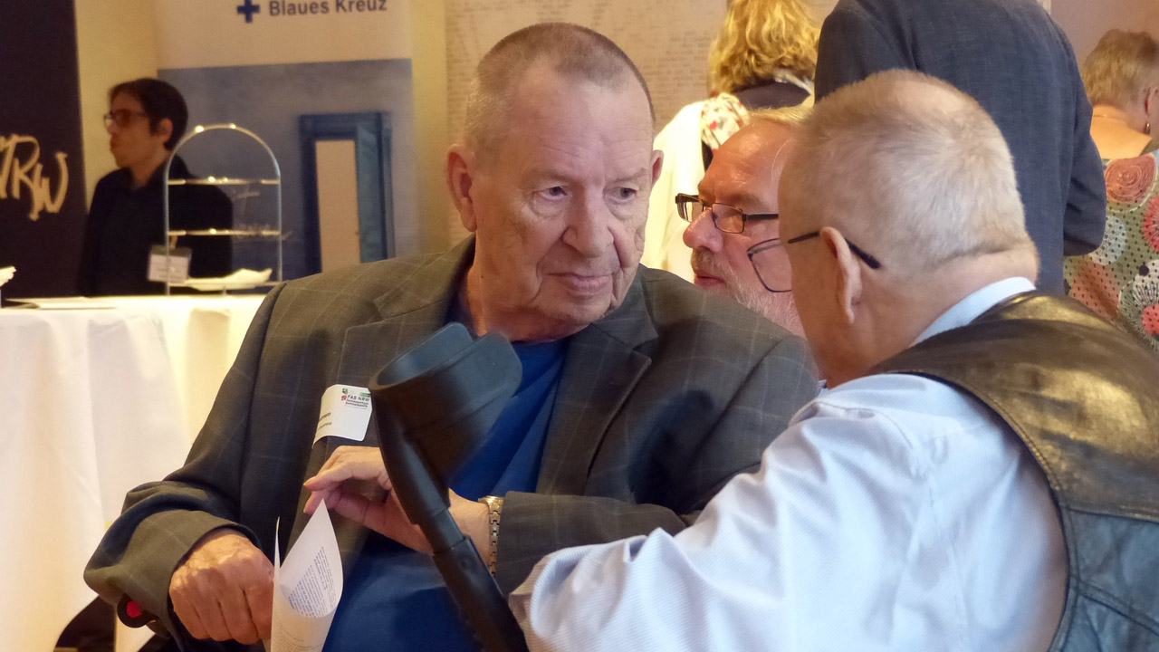 Zwei ältere Männer im Gespräch auf einer Veranstaltung: Ein Mann mit grauem Haar in kariertem Sakko und blauem Shirt hält eine Unterarmgehstütze und ein gefaltetes Blatt Papier; ein zweiter Mann mit weißem Haar, Brille, hellblauem Hemd und schwarzer Lederweste beugt sich zu ihm. Im Hintergrund weitere Gäste, ein Stehtisch mit Etagere und ein Banner mit der Aufschrift 'Blaues Kreuz'.