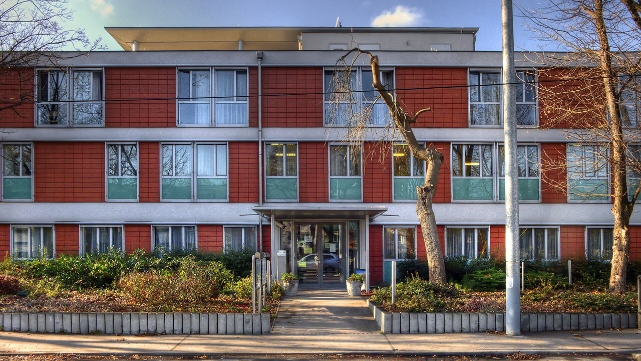 Eingangsbereich des Anton-Proksch-Instituts mit roter Klinkerfassade und weißen horizontalen Bändern an einem sonnigen Tag. Das Haus hat regelmäßige Fensterreihen und einen mittigen, überdachten Eingang. Davor befinden sich niedrige Hecken, ein gepflasterter Weg und ein kahler Baum.