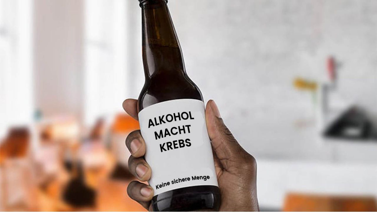 Nahaufnahme einer Hand, die eine braune Bierflasche gegen einen unscharfen Innenhintergrund hält. Das weiße Etikett der Flasche trägt in schwarzer Großschrift den warnenden Text „Alkohol macht Krebs“ sowie darunter in kleinerer Schrift „Keine sichere Menge“.
