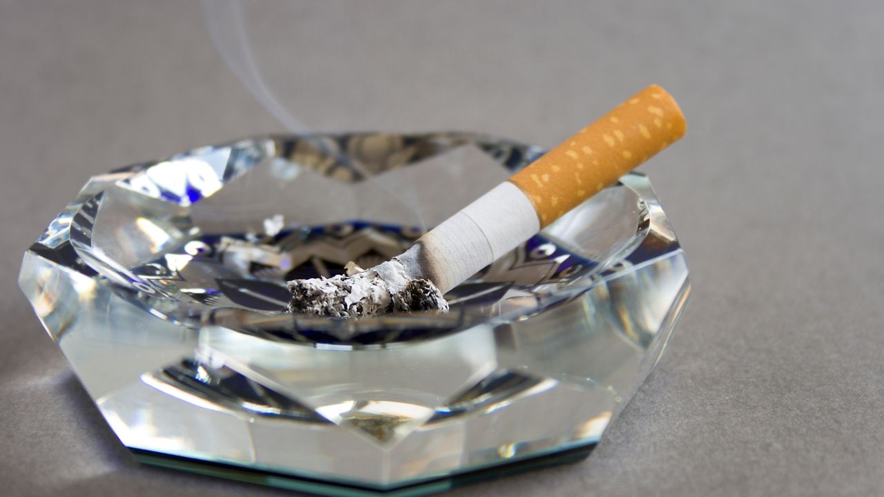 Nahaufnahme einer brennenden Zigarette mit orangefarbenem Filter, die in einem geschliffenen Kristall-Aschenbecher liegt; Asche und feiner Rauch sind sichtbar.