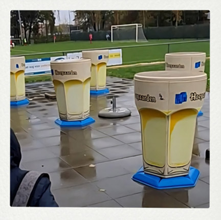 Mehrere Stehtische in Form von Hoegaarden-Biergläsern stehen auf einem nassen, gepflasterten Vorplatz neben einem Fußballfeld.
