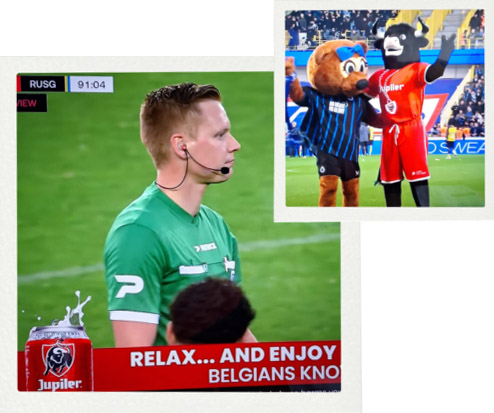 Geteiltes TV-Bild von einem Fußballspiel. Das große Bild links zeigt einen Schiedsrichter in grünem Trikot mit Headset-Mikrofon auf dem Rasen. Oben links die Spielanzeige 'RUSG 91:04'. Unten ein rotes Werbebanner von Jupiler-Bier mit dem Text 'RELAX... AND ENJOY BELGIANS KNOWHOW'. Das kleine Bild rechts oben zeigt zwei Fußball-Maskottchen auf dem Platz die winken: links ein brauner Bär mit blauer Schleife im blau-schwarz gestreiften Trikot, rechts ein schwarzer Stier im roten Trikot.