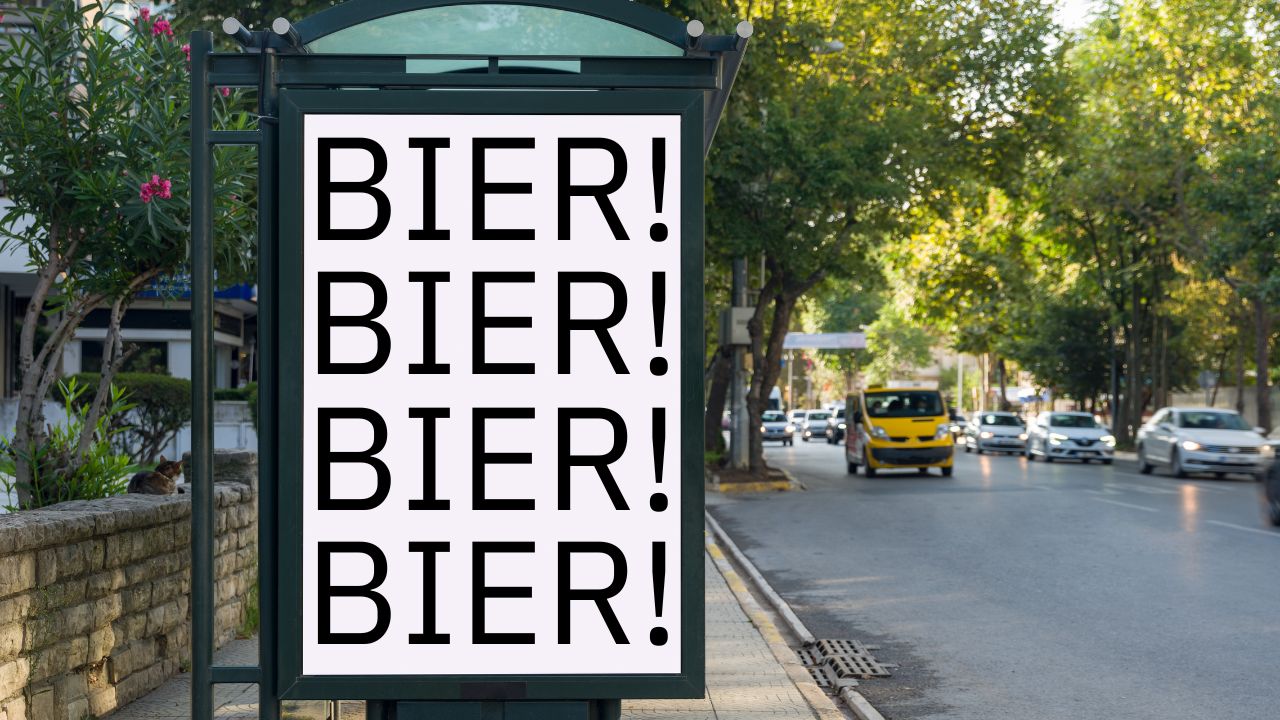Vierfach wiederholtes 'BIER!'-Plakat in großen schwarzen Buchstaben an einer Bushaltestelle. Sonnige Straße mit Autos und Bäumen im Hintergrund, links eine Katze auf einer Mauer.