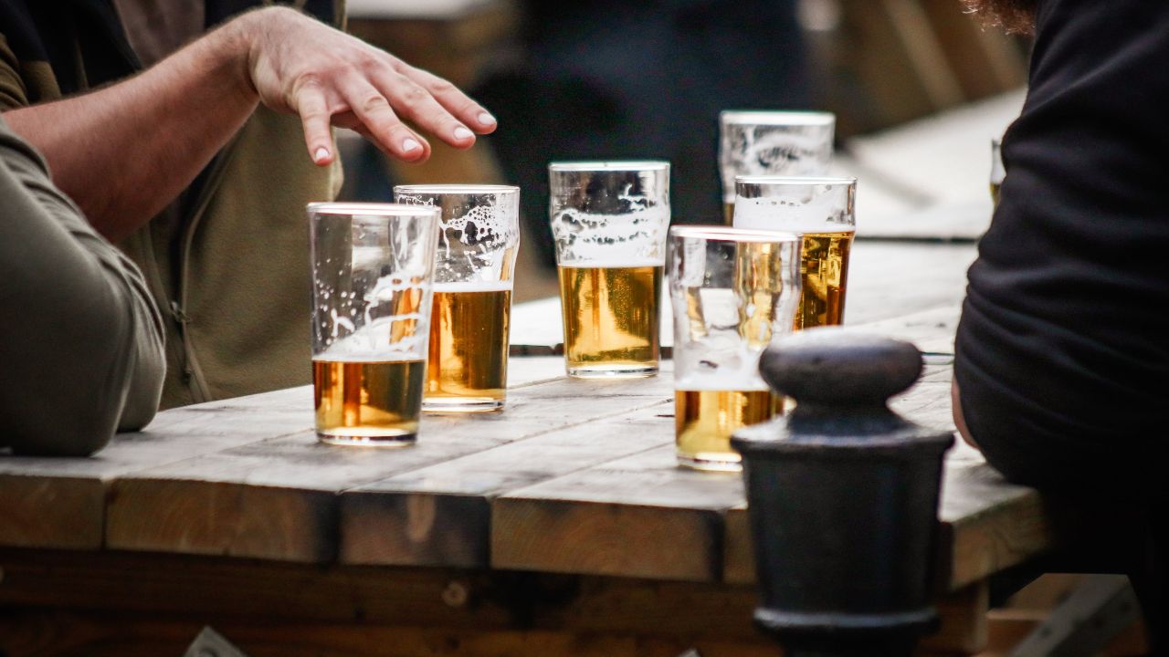Mehrere halbvolle Biergläser auf einem rustikalen Holztisch, um den sich im Freien sitzende Personen unterhalten.