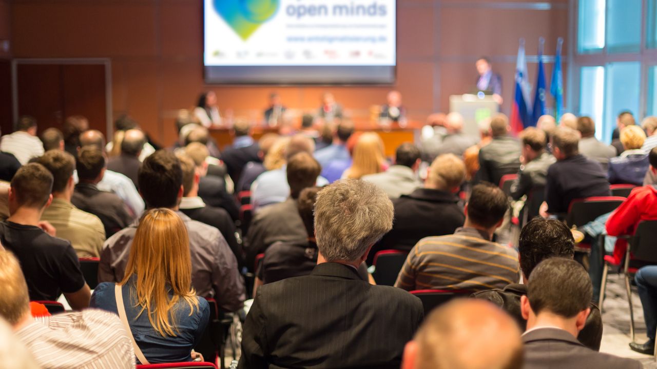 Publikum von hinten bei einer Konferenz: Zahlreiche Zuhörer*innen sitzen in Reihen und blicken auf eine Bühne, auf der ein Redner am Pult steht. Im Hintergrund sind eine Leinwand mit dem Schriftzug 'open minds' sowie mehrere Flaggen zu sehen.