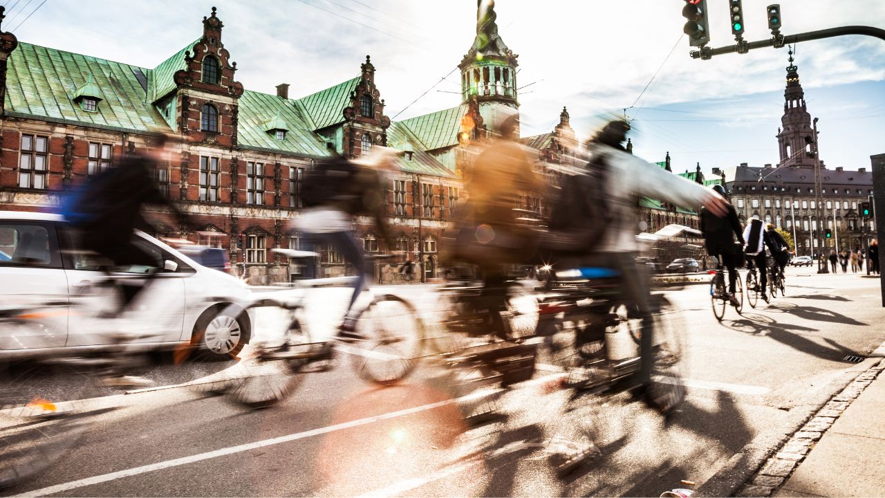 Mehrere Radfahrer*innen fahren mit Bewegungsunschärfe vor einem historischen Gebäude im Zentrum Kopenhagens über eine Kreuzung.