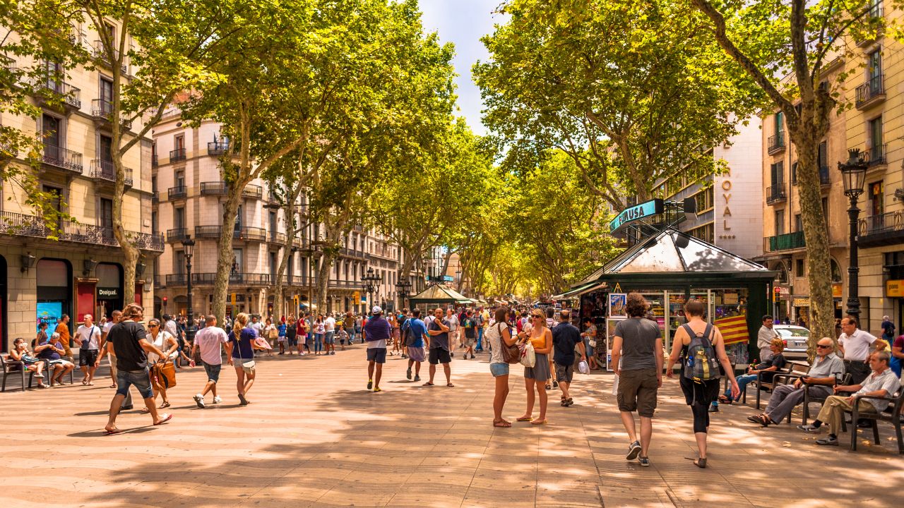 Belebte Flaniermeile La Rambla in Barcelona an einem sonnigen Tag mit zahlreichen Passanten, Platanen, die Schatten spenden, historischen Gebäudefassaden mit Balkonen auf beiden Seiten, Zeitungskiosken und dem Hotel Royal im Hintergrund.