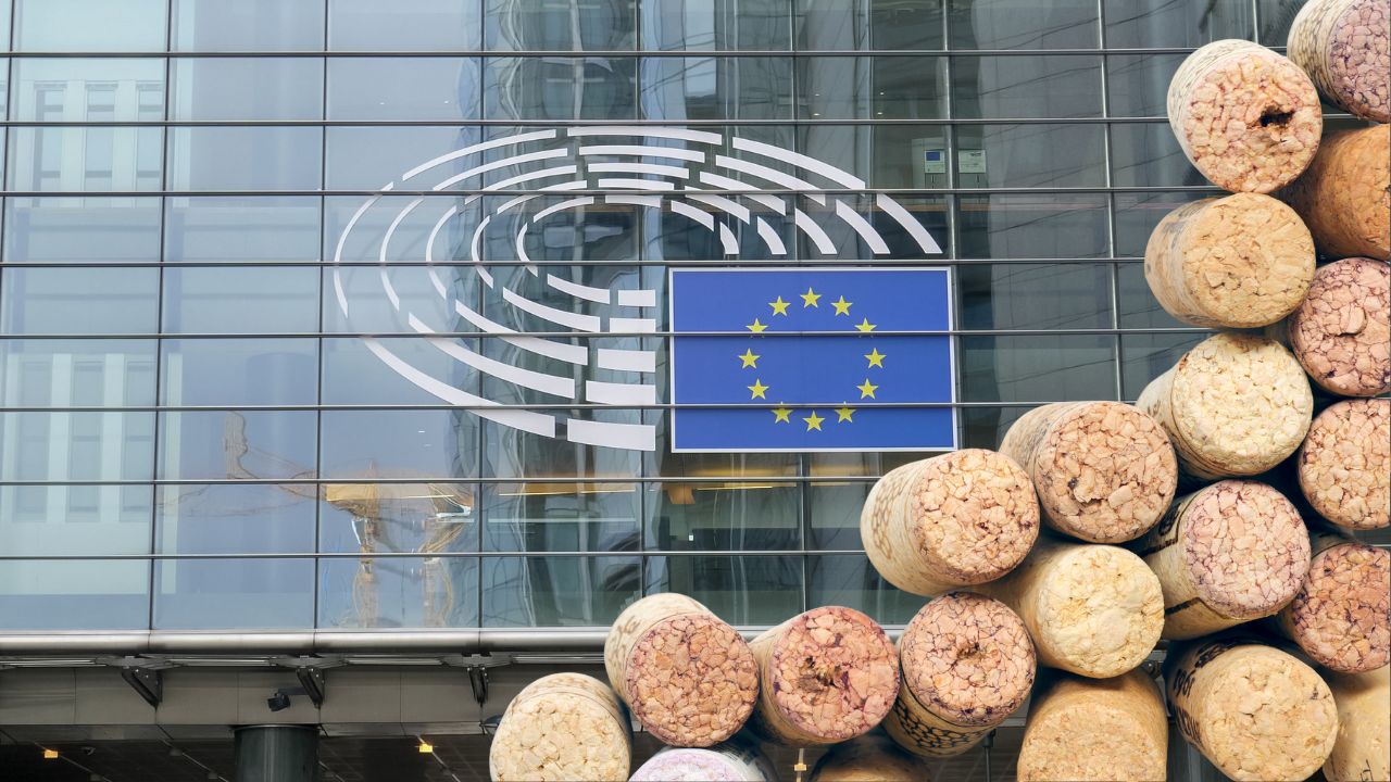 Außenansicht des Europäischen Parlaments mit dem Logo aus blauer Flagge und stilisiertem Plenarsaal auf der Glasfront, davor stapeln sich zahlreiche gebrauchte Weinkorken.