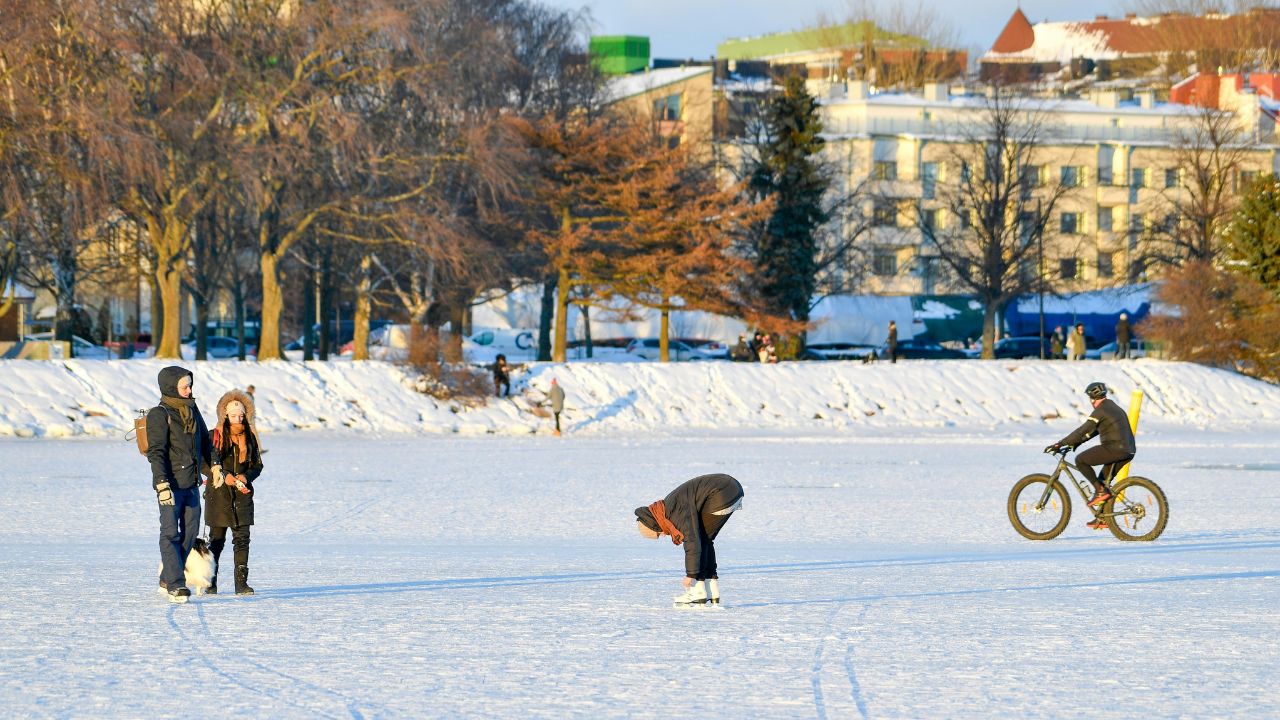 Winterlandschaft mit zugefrorenem Gewässer, auf dem mehrere Personen aktiv sind: Links steht ein Paar mit einem weißen Hund, in der Mitte beugt sich eine Person beim Eislaufen nach vorne, rechts fährt ein Radfahrer mit Helm über das Eis. Im Hintergrund verschneite Uferböschungen, kahle Bäume und Gebäude bei sonnigem Wetter.