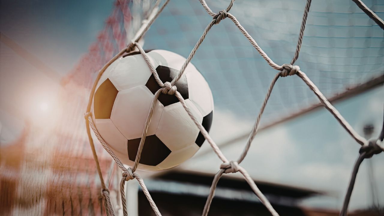 Nahaufnahme eines schwarz-weißen Fußballs, der sich in die Maschen eines Tornetzes drückt, mit unscharfem blauen Himmel und Stadion im Hintergrund.